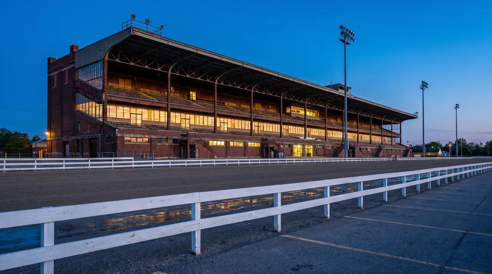 Generated image: a long brick and steel grandstand at blue hour with three floors of lit windows, seen across an empty racetrack and a white rail; not a photograph of the casino building.